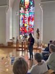 Auf dem Foto sitzen Menschen am rechten Rand in einer Kapelle. Eine Person steht in Richtung Altar. Im Hintergrund scheint Licht durch ein buntes, großes Kirchenfenster. Auf dem Boden liegt ein Fischernetz und darauf Fische aus Papier.
